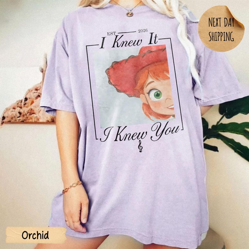 May include: A light purple short-sleeve t-shirt featuring a graphic print of a cartoon character with red hair and a red hat. The text reads EST 2026 I Knew It I Knew You with a musical note symbol. The design is framed by a thin black rectangular border.