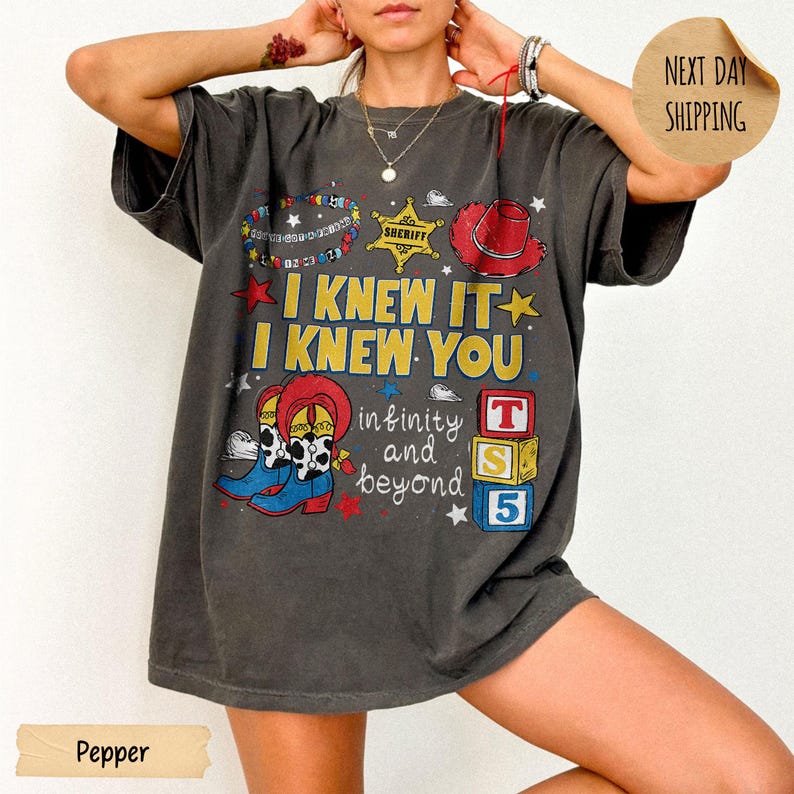 May include: A charcoal gray short-sleeve t-shirt featuring a graphic print with the text I KNEW IT I KNEW YOU infinity and beyond. The design includes a sheriff badge, a red cowboy hat, blue cowboy boots, and three stacked blocks labeled T S 5.