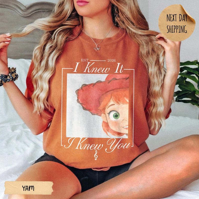 May include: A burnt orange crewneck sweatshirt featuring a graphic print of a cartoon character with red hair and a red hat. The text reads EST 2026 I Knew It I Knew You with a small musical note symbol. The shirt has a relaxed fit and short sleeves.