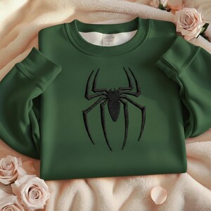 Puede incluir: Una sudadera verde oscuro con un dise&ntilde;o de ara&ntilde;a negra bordado en la parte delantera. La sudadera tiene cuello redondo y mangas largas. El dise&ntilde;o de la ara&ntilde;a est&aacute; centrado en el pecho.
