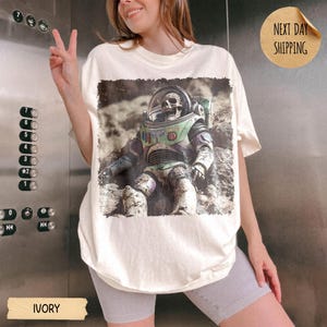 May include: An ivory t-shirt featuring a graphic print of a skeleton wearing a space suit with a clear helmet. The design shows the figure sitting on a rocky, cratered surface. The shirt has a crew neckline and short sleeves, with a loose fit.