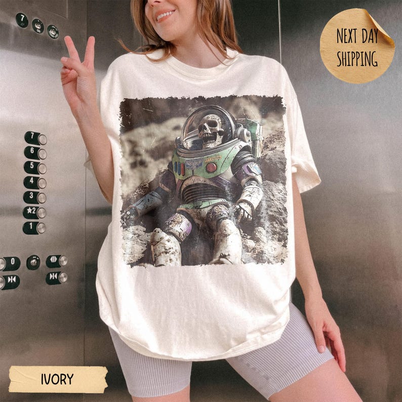May include: An ivory t-shirt featuring a graphic print of a skeleton wearing a space suit with a clear helmet. The design shows the figure sitting on a rocky, cratered surface. The shirt has a crew neckline and short sleeves, with a loose fit.