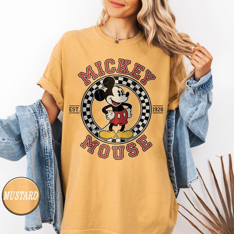 Puede incluir: Camiseta de color mostaza con un gr&aacute;fico vintage de Mickey Mouse. El dise&ntilde;o incluye las palabras "Mickey Mouse" en un arreglo circular, con "Est. 1928" y un fondo a cuadros. La camiseta tiene mangas enrolladas.