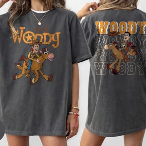 May include: Dark gray t-shirt featuring a cartoon image of Woody riding a horse, with the text "Woody" in an orange and yellow font. The back of the shirt repeats the image and text.
