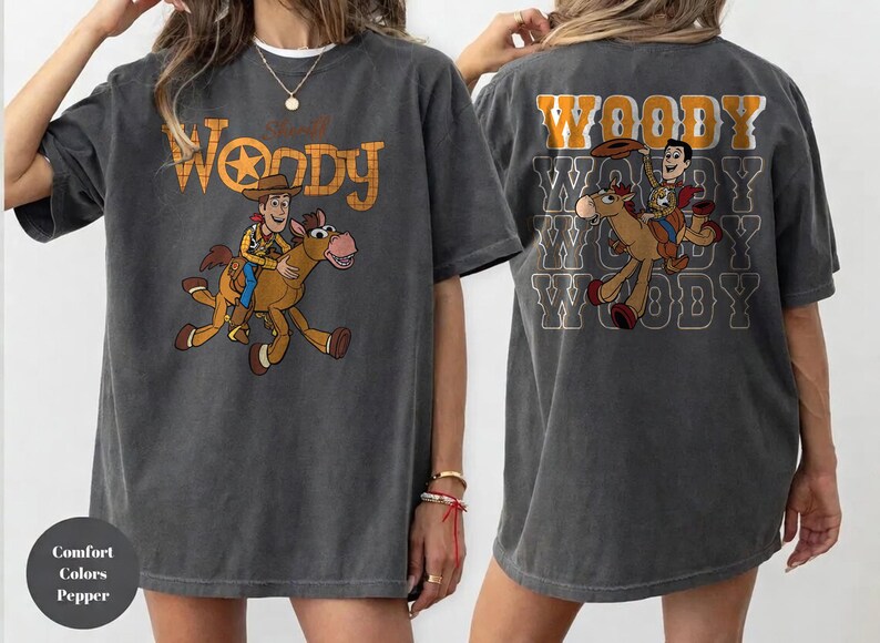 May include: Dark gray t-shirt featuring a cartoon image of Woody riding a horse, with the text "Woody" in an orange and yellow font. The back of the shirt repeats the image and text.