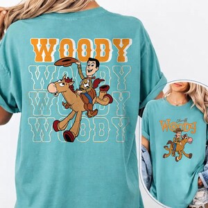 May include: A turquoise t-shirt featuring a graphic of Woody from Toy Story riding a horse. The word "WOODY" is printed in orange and white block letters above the graphic. The shirt has a relaxed fit.