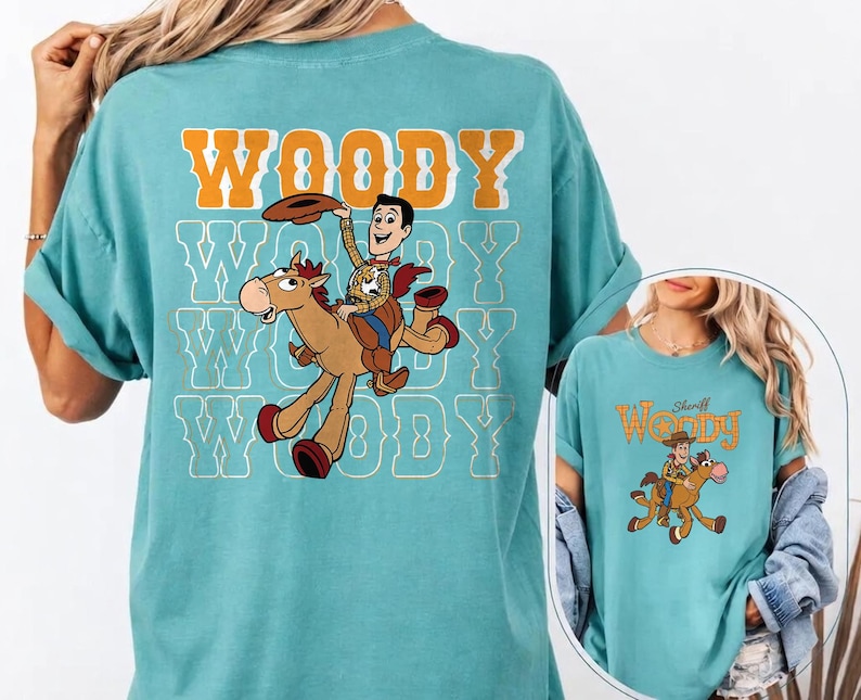 May include: A turquoise t-shirt featuring a graphic of Woody from Toy Story riding a horse. The word "WOODY" is printed in orange and white block letters above the graphic. The shirt has a relaxed fit.