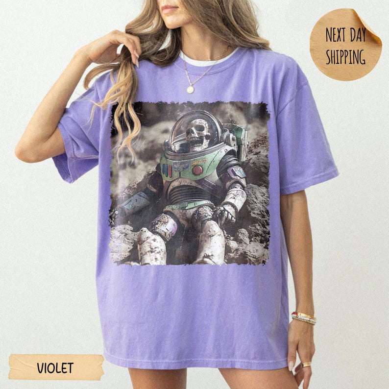 May include: A violet short-sleeve t-shirt featuring a graphic print of a skeleton wearing a space suit with a clear helmet. The astronaut figure is positioned on a rocky, cratered surface. The shirt has a crew neckline and a loose fit.