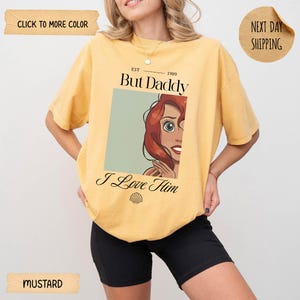 Op de afbeelding: Een mosterdgeel T-shirt met korte mouwen en een grafische print van een personage met rood haar. De tekst luidt EST 1989 But Daddy I Love Him met een klein schelppictogram eronder. Het ontwerp staat centraal op de borst van het kledingstuk.