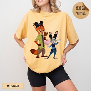 May include: Mustard-colored t-shirt featuring a cartoon illustration of Nick Wilde and Judy Hopps from Zootopia. Nick is wearing a green shirt and tie, Judy is in a police uniform. The shirt has a relaxed fit.