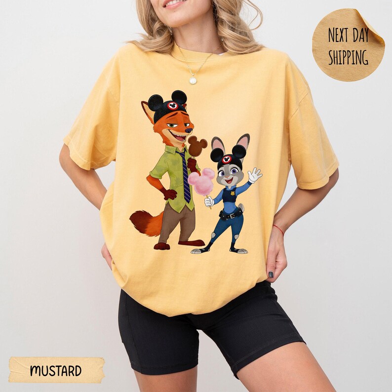 May include: Mustard-colored t-shirt featuring a cartoon illustration of Nick Wilde and Judy Hopps from Zootopia. Nick is wearing a green shirt and tie, Judy is in a police uniform. The shirt has a relaxed fit.