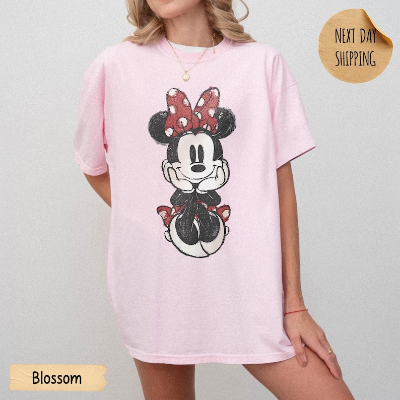 May include: A light pink short-sleeve t-shirt featuring a distressed graphic print of a cartoon mouse with a red and white polka dot bow. The mouse is depicted with hands resting under its chin. The shirt has a crew neckline and a relaxed fit.