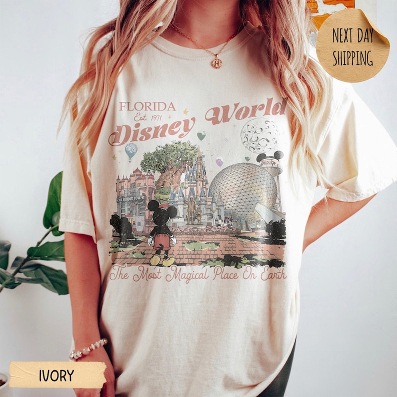 May include: Ivory-colored t-shirt featuring a vintage-style graphic of Disney World in Florida. The design includes Mickey Mouse, a castle, Spaceship Earth, and the text "Florida Disney World Est. 1971" and "The Most Magical Place On Earth."