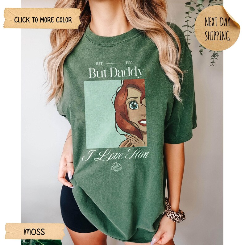 Op de afbeelding: Een mosgroen t-shirt met korte mouwen en een grafische print van een roodharig personage in een vierkant kader. De tekst luidt EST 1989, But Daddy, en I Love Him, vergezeld door een klein schelppictogram onder de afbeelding.