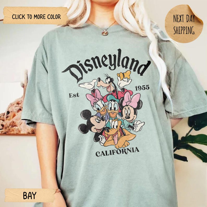 Op de afbeelding: Saliegroen t-shirt met een afbeelding van Disney-personages, waaronder Mickey Mouse, Minnie Mouse, Donald Duck en Goofy. Het woord "Disneyland" staat in een boog bovenaan, met "California" onderaan. Het shirt heeft ook de tekst "Est 1955".