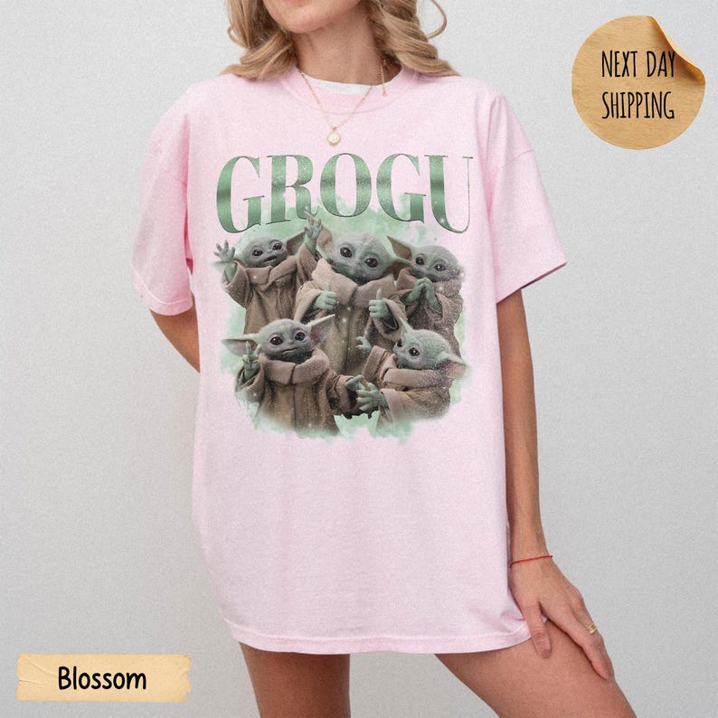May include: A light pink short-sleeve t-shirt featuring a graphic print of five small green creatures in tan robes. The text GROGU is printed in large green letters above the creatures. The shirt has a crew neckline and a relaxed fit.