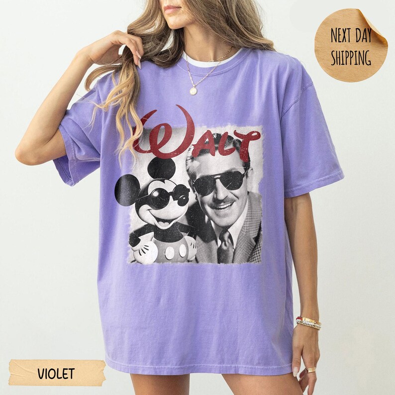 May include: Violet-colored t-shirt featuring a black and white graphic of Mickey Mouse and Walt Disney. The word "WALT" is printed in red above the image. The shirt has short sleeves and a crew neck.