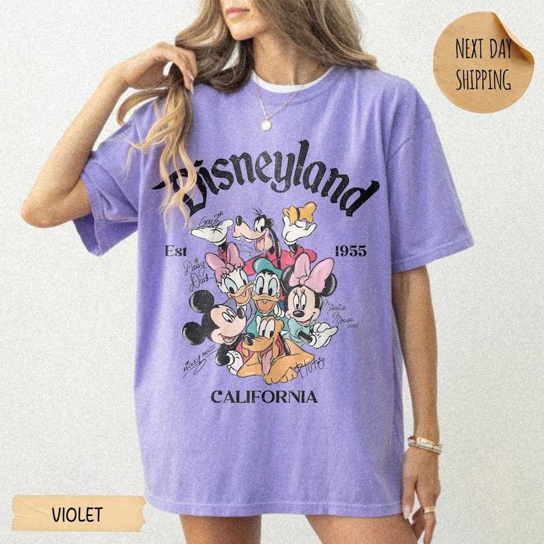 May include: A violet short-sleeve t-shirt featuring a graphic print of Disney characters including Mickey Mouse, Minnie Mouse, Donald Duck, Daisy Duck, Goofy, and Pluto. The text reads Disneyland, Est 1955, and California in black lettering.