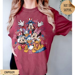 May include: A crimson t-shirt featuring a graphic print of Disney characters including Mickey Mouse, Donald Duck, Goofy, Daisy Duck, Minnie Mouse, and Pluto. The design includes American flags, fireworks, and text reading America 250th Anniversary 1776-2026.