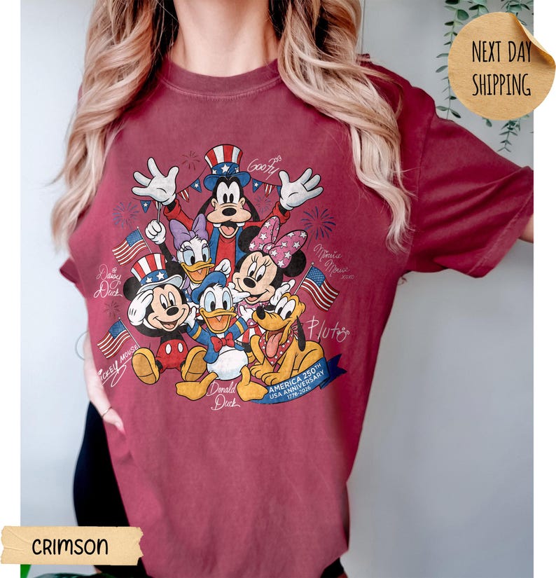 May include: A crimson t-shirt featuring a graphic print of Disney characters including Mickey Mouse, Donald Duck, Goofy, Daisy Duck, Minnie Mouse, and Pluto. The design includes American flags, fireworks, and text reading America 250th Anniversary 1776-2026.