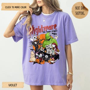 May include: A violet short-sleeve t-shirt featuring a graphic print with the text Nightmare on Main Street. The design includes characters from The Nightmare Before Christmas, such as Jack Skellington, Sally, and Oogie Boogie, set against an orange moon.