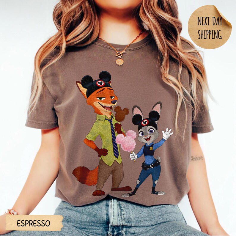 May include: Espresso-colored t-shirt featuring a cartoon fox and bunny wearing Mickey Mouse ears. The fox is in a green shirt and brown pants, while the bunny is in a police uniform. The shirt also has the text "Next Day Shipping".