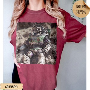 May include: A crimson t-shirt featuring a graphic print of a skeleton wearing a space suit with a clear helmet, set against a rocky lunar landscape. The design includes green, purple, and white accents on the suit, with a weathered, textured appearance.