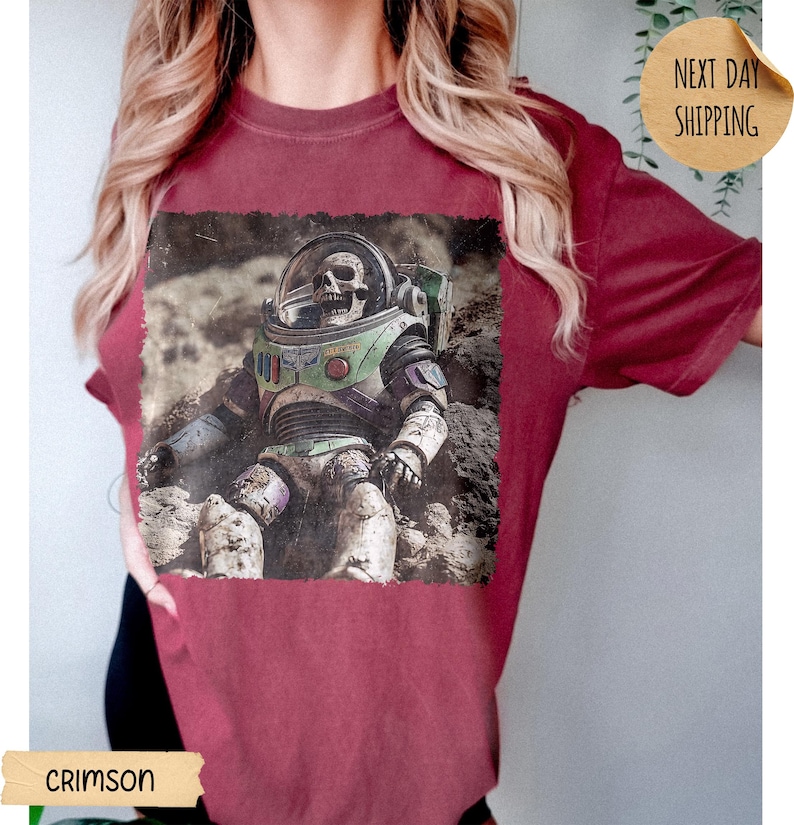 May include: A crimson t-shirt featuring a graphic print of a skeleton wearing a space suit with a clear helmet, set against a rocky lunar landscape. The design includes green, purple, and white accents on the suit, with a weathered, textured appearance.