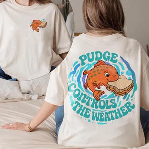 May include: Off-white t-shirt with a cartoon orange fish and sandwich graphic. The back of the shirt has the text "Pudge Controls The Weather" in teal and blue. A smaller version of the fish and sandwich is on the front.