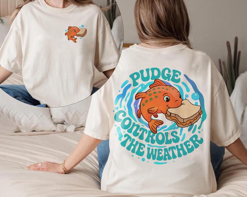 May include: Off-white t-shirt with a cartoon orange fish and sandwich graphic. The back of the shirt has the text "Pudge Controls The Weather" in teal and blue. A smaller version of the fish and sandwich is on the front.