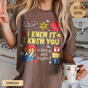 May include: A brown t-shirt featuring a graphic print with the text I KNEW IT I KNEW YOU and infinity and beyond. The design includes a beaded bracelet, a yellow sheriff star, a red cowboy hat, cowboy boots, and three stacked alphabet blocks.