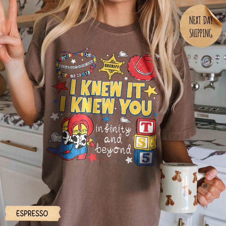 May include: A brown t-shirt featuring a graphic print with the text I KNEW IT I KNEW YOU and infinity and beyond. The design includes a beaded bracelet, a yellow sheriff star, a red cowboy hat, cowboy boots, and three stacked alphabet blocks.