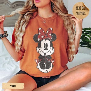 May include: A burnt orange crewneck t-shirt featuring a distressed graphic of Minnie Mouse with a red and white polka dot bow, hands resting under the chin. The shirt has a relaxed fit and a vintage-style print of the character in black and white.