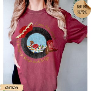 May include: A crimson short-sleeve t-shirt featuring a graphic of a black tire framing a scene with three cartoon characters. The design includes a red cowboy hat, a red and white cowboy boot, and the text Love has ways of bringing things back to life.