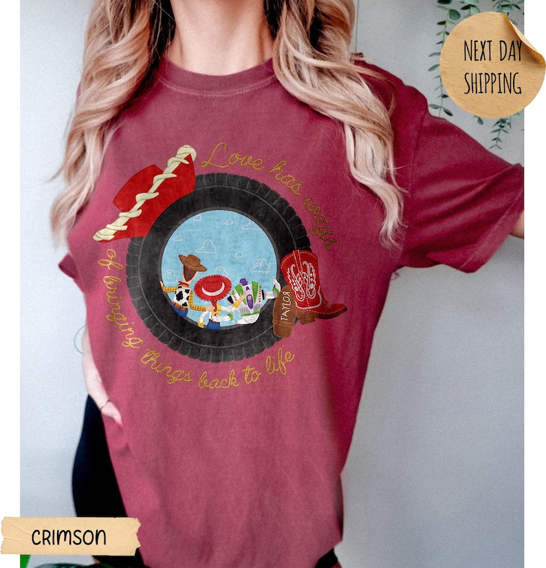 May include: A crimson short-sleeve t-shirt featuring a graphic of a black tire framing a scene with three cartoon characters. The design includes a red cowboy hat, a red and white cowboy boot, and the text Love has ways of bringing things back to life.