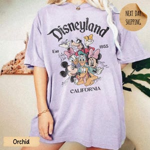 May include: A light purple short-sleeve t-shirt featuring a graphic print of Disney characters including Mickey Mouse, Minnie Mouse, Donald Duck, Daisy Duck, Goofy, and Pluto. The text reads Disneyland Est 1955 California.