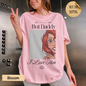 Op de afbeelding: Een lichtroze t-shirt met een afbeelding van een roodharig personage, de tekst "EST 1989 But Daddy I Love Him" en een klein schelppictogram. Het ontwerp is gecentreerd op de borst van het kledingstuk met korte mouwen en een ronde hals.