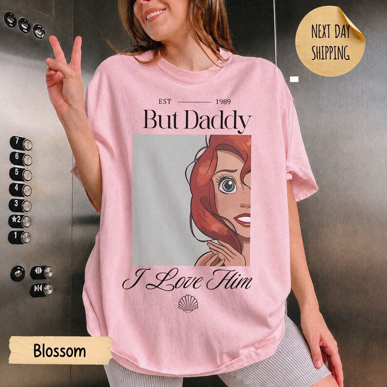 Op de afbeelding: Een lichtroze t-shirt met een afbeelding van een roodharig personage, de tekst "EST 1989 But Daddy I Love Him" en een klein schelppictogram. Het ontwerp is gecentreerd op de borst van het kledingstuk met korte mouwen en een ronde hals.