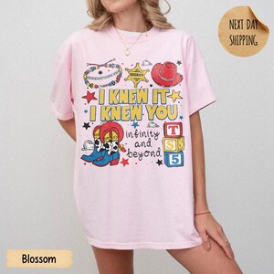 May include: A light pink t-shirt featuring a graphic print with the text I KNEW IT I KNEW YOU infinity and beyond. The design includes a beaded bracelet, a sheriff star, a red cowboy hat, cowboy boots, and three colorful alphabet blocks.