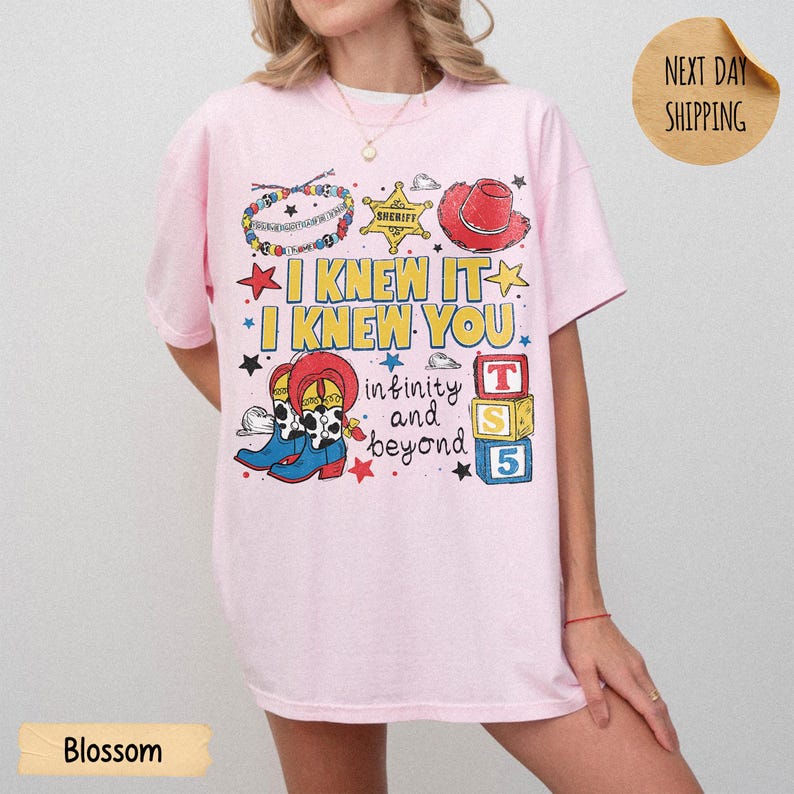 May include: A light pink t-shirt featuring a graphic print with the text I KNEW IT I KNEW YOU infinity and beyond. The design includes a beaded bracelet, a sheriff star, a red cowboy hat, cowboy boots, and three colorful alphabet blocks.