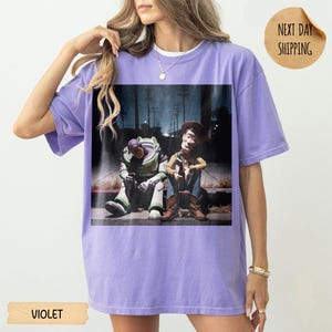 Pode incluir: Camiseta violeta com um gr&aacute;fico de Buzz Lightyear e Woody de Toy Story. Os personagens s&atilde;o retratados sentados. A camiseta tem gola redonda e mangas curtas. O texto "VIOLET" est&aacute; na parte inferior.
