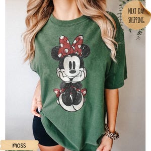 May include: A moss green short-sleeve t-shirt featuring a distressed graphic print of Minnie Mouse with her hands under her chin, wearing a red and white polka dot bow. The shirt has a crew neckline and a relaxed, loose-fitting silhouette.