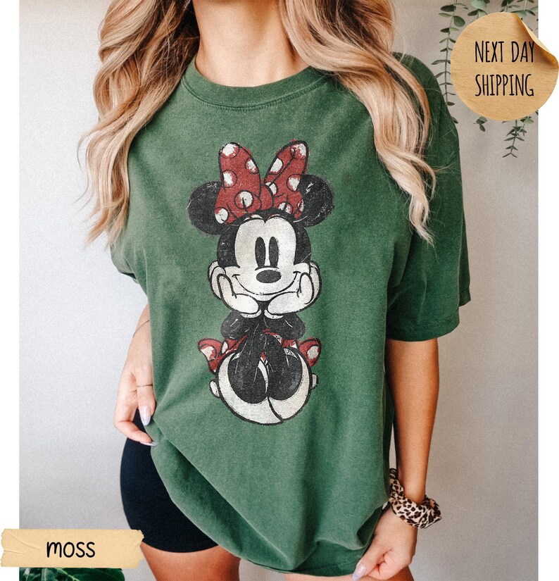 May include: A moss green short-sleeve t-shirt featuring a distressed graphic print of Minnie Mouse with her hands under her chin, wearing a red and white polka dot bow. The shirt has a crew neckline and a relaxed, loose-fitting silhouette.