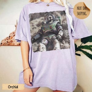 May include: A light purple short-sleeve t-shirt featuring a rectangular graphic print of a skeleton wearing a space suit with a green chest plate and a clear helmet, depicted sitting on a rocky, dusty surface resembling the moon's terrain.