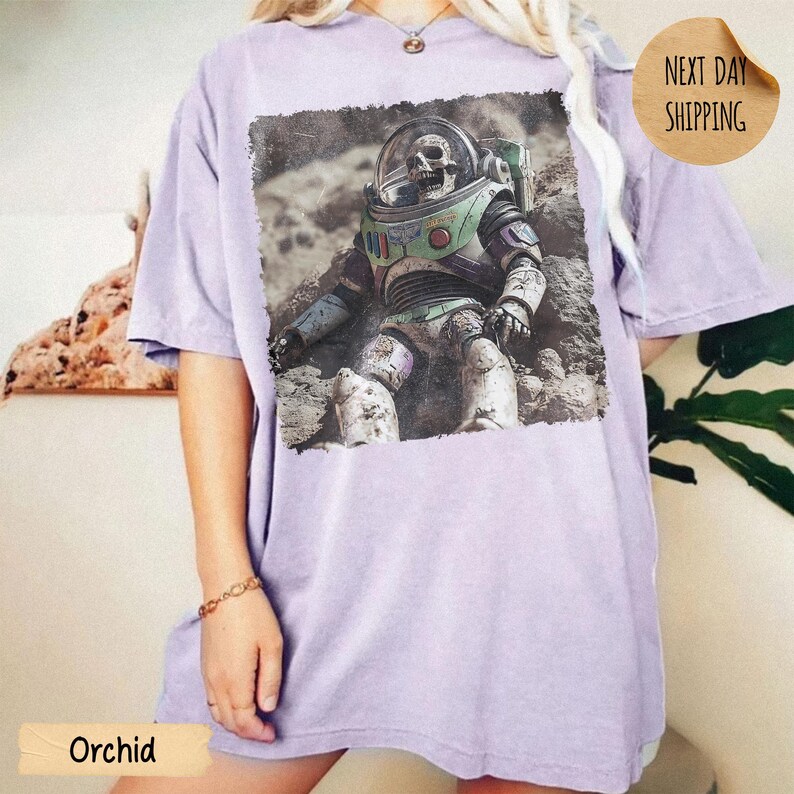 May include: A light purple short-sleeve t-shirt featuring a rectangular graphic print of a skeleton wearing a space suit with a green chest plate and a clear helmet, depicted sitting on a rocky, dusty surface resembling the moon's terrain.