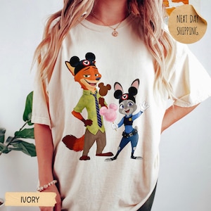 May include: Ivory-colored t-shirt featuring a cartoon fox and bunny with Mickey Mouse ears, holding treats. The fox wears a green shirt and brown pants, while the bunny is in a police uniform. The shirt has the text "IVORY" and "NEXT DAY SHIPPING".