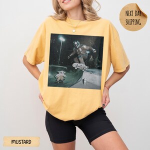 May include: Mustard yellow t-shirt featuring a graphic of a skater and Grogu from The Mandalorian. The image shows the skater in action at a skate park. The shirt has a relaxed fit.
