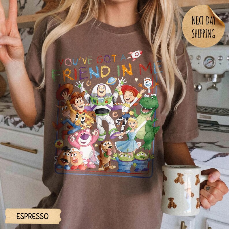 May include: A brown short-sleeve t-shirt featuring a colorful graphic print of various animated characters from the Toy Story franchise. The text "YOU'VE GOT A FRIEND IN ME" is printed above the characters in a multi-colored, playful font style.