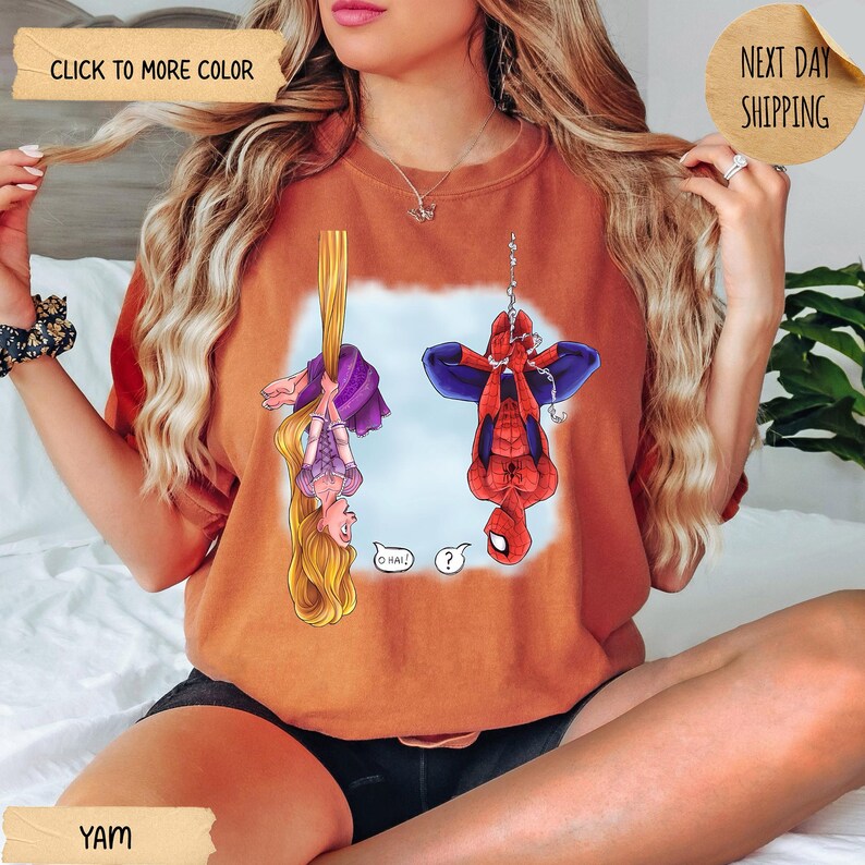 Op de afbeelding: Een roestoranje sweater met ronde hals en een grafische print van een blond personage met lang haar dat aan een vlecht hangt en een rood-blauw superheldpersonage dat aan een web hangt. De personages kijken elkaar aan met tekstballonnen.