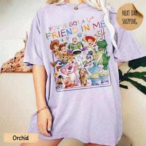 May include: A light purple t-shirt featuring a colorful graphic print of various animated characters from the Toy Story franchise. The text "YOU'VE GOT A FRIEND IN ME" is printed in multi-colored letters above the group of characters.